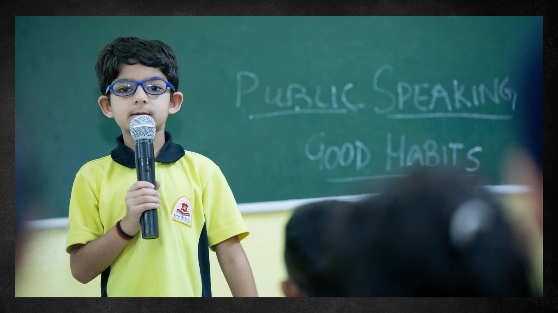 A young student holding a microphone and speaking in front of the class during a public speaking activity.