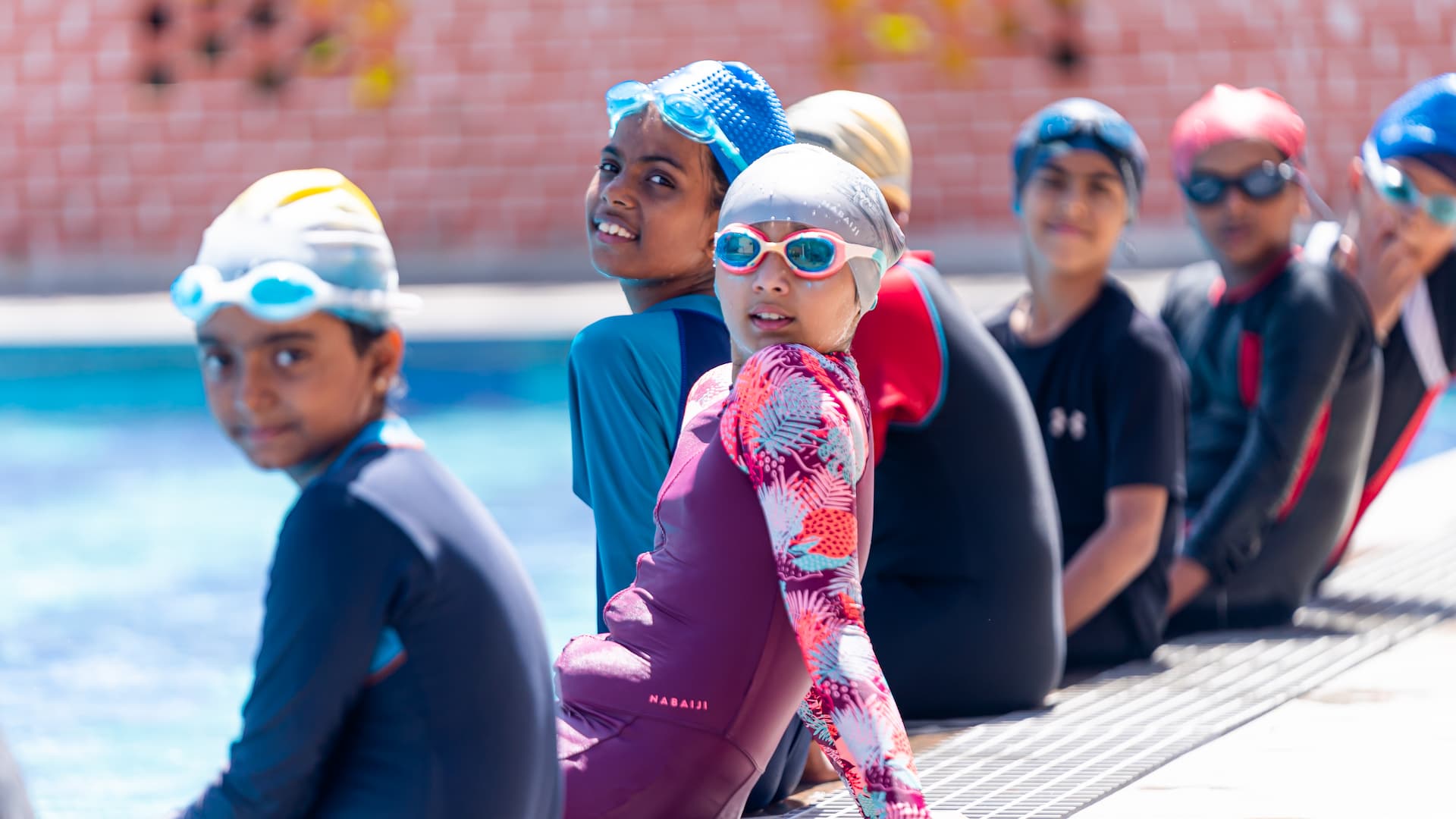 School kids swimming in the pool while following safety rules, wearing proper swimwear, caps, & goggles, as teachers guide them through the lesson.