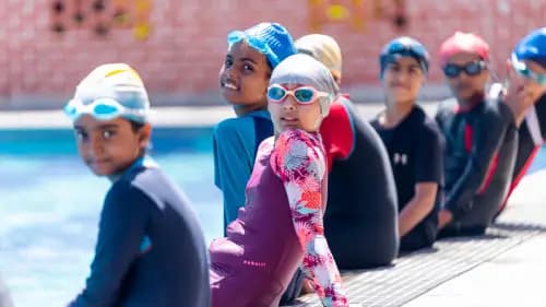 School kids swimming in the pool while following safety rules, wearing proper swimwear, caps, & goggles, as teachers guide them through the lesson.