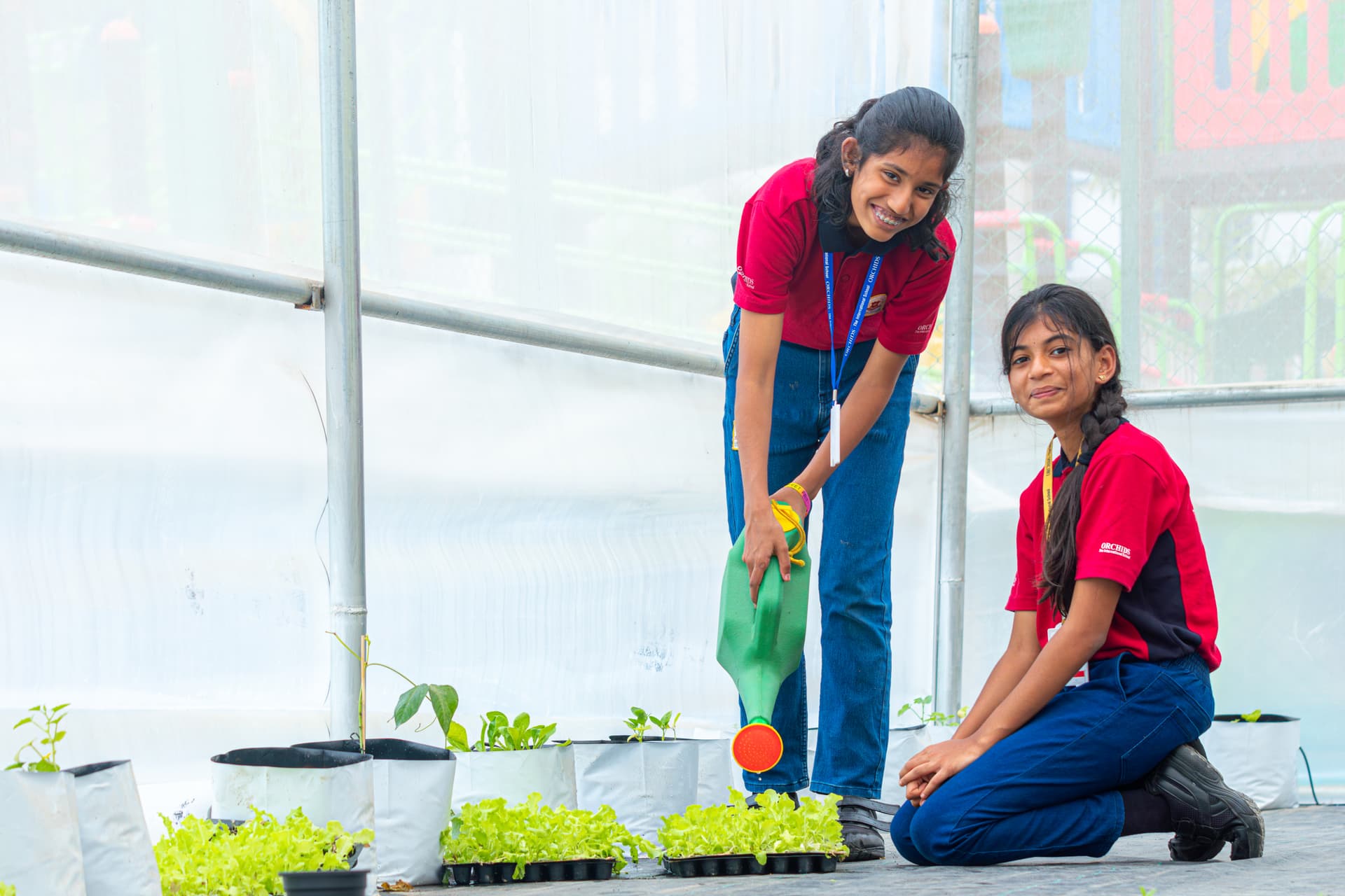 Two girls in red school uniforms engage in a horticulture activity, smiling as they water and care for plants inside a greenhouse.